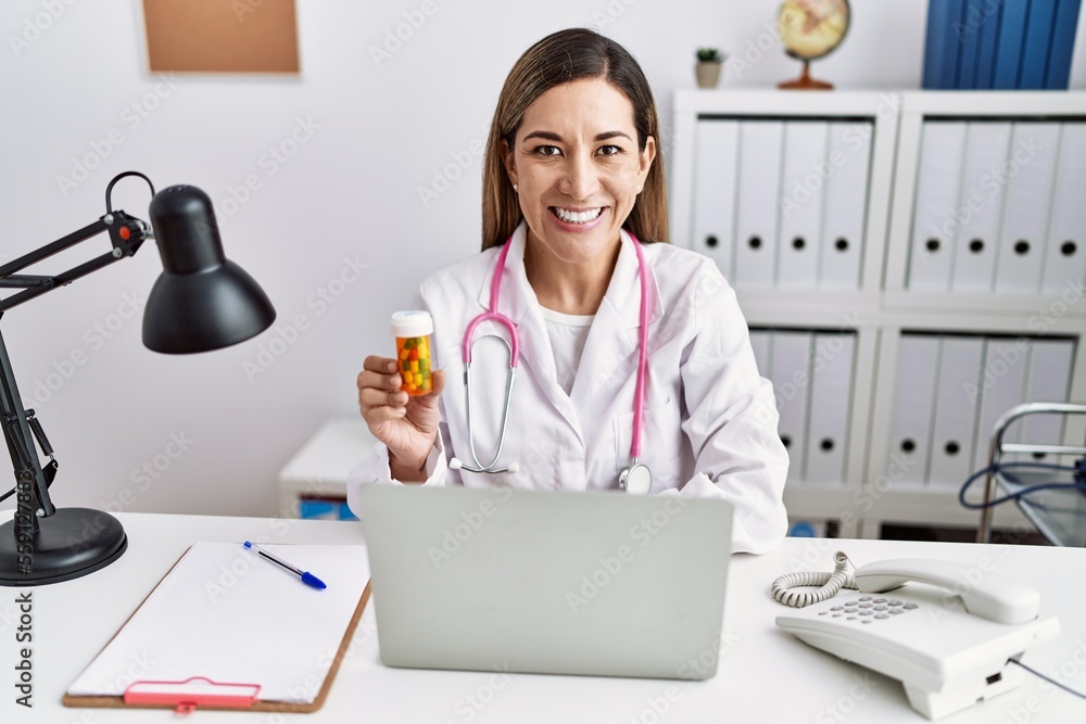Young hispanic woman wearing doctor uniform holding pills at the clinic with a happy and cool smile on face. lucky person. Young hispanic woman wearing doctor uniform holding pills at the clinic with a happy and cool smile on face. lucky person.
