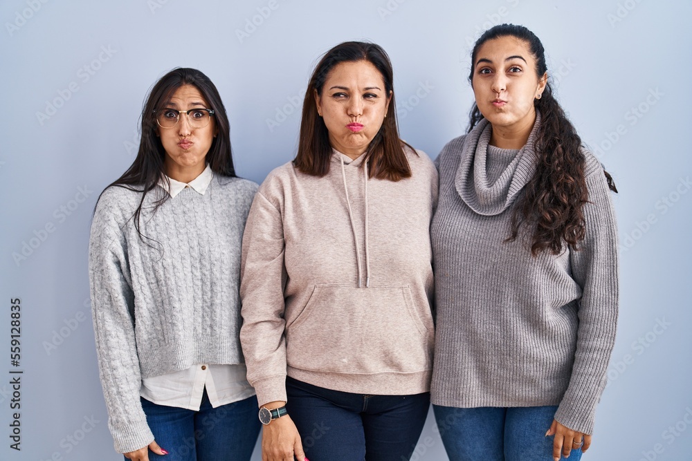 Mother and two daughters standing over blue background puffing cheeks with funny face. mouth inflated with air, crazy expression. - obrazy, fototapety, plakaty 