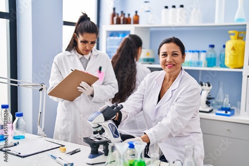 Three woman scientists using microscope write on checklist at laboratory