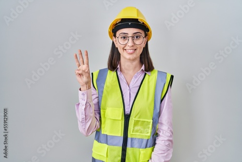 Hispanic girl wearing builder uniform and hardhat showing and pointing up with fingers number three while smiling confident and happy.
