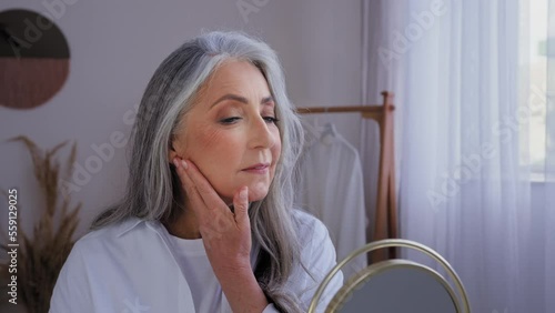 Smiling beautiful aging 50s yeas old lady skin care at home moisture looking at mirror reflection touch face cream facial anti-wrinkled cosmetic 60s senior mature woman touching cheeks lifting wrinkle photo