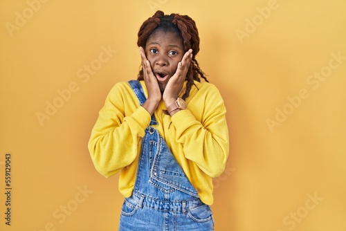 African woman standing over yellow background afraid and shocked, surprise and amazed expression with hands on face