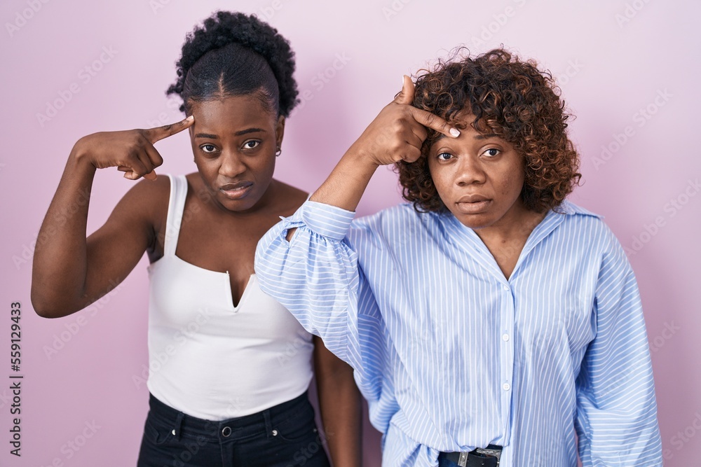 Two african women standing over pink background pointing unhappy to