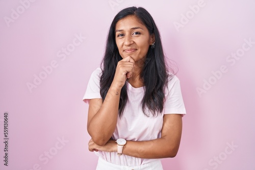 Young hispanic woman standing over pink background with hand on chin thinking about question  pensive expression. smiling and thoughtful face. doubt concept.