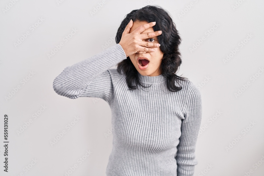 Hispanic woman with dark hair standing over isolated background peeking in shock covering face and eyes with hand, looking through fingers with embarrassed expression. Hispanic woman with dark hair standing over isolated background peeking in shock covering face and eyes with hand, looking through fingers with embarrassed expression.