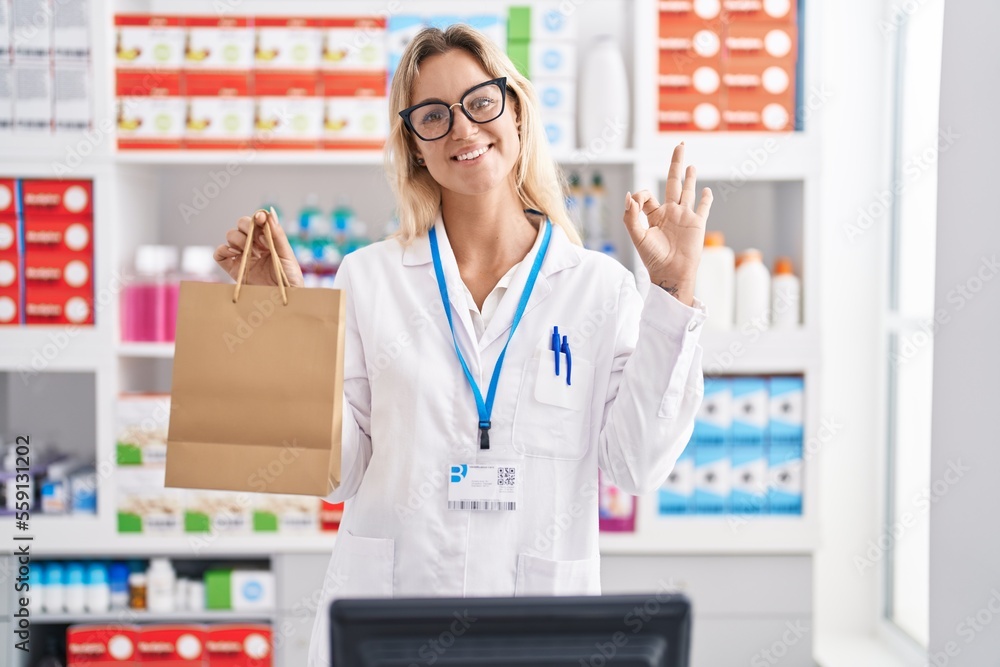 Young blonde woman working at pharmacy drugstore holding paper bag doing ok sign with fingers, smiling friendly gesturing excellent symbol