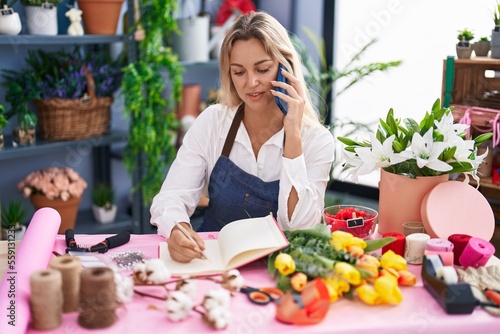 Young blonde woman florist talking on smartphone writing on notebook at florist