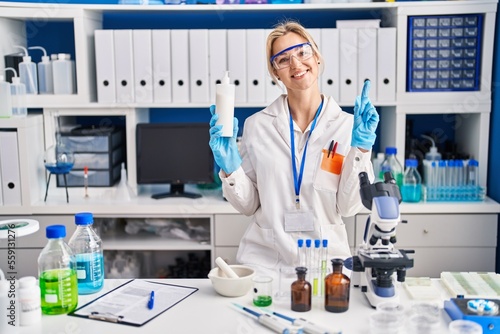 Young caucasian woman working at scientist laboratory holding body lotion smiling with an idea or question pointing finger with happy face  number one