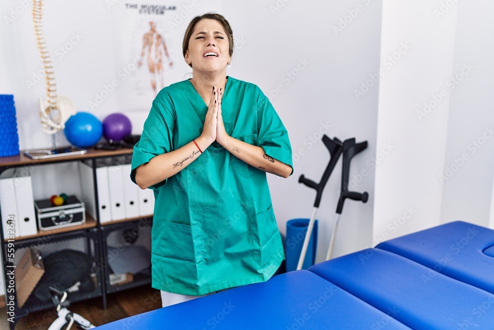 Young hispanic woman wearing physiotherapist uniform standing at clinic begging and praying with hands together with hope expression on face very emotional and worried. begging. Young hispanic woman wearing physiotherapist uniform standing at clinic begging and praying with hands together with hope expression on face very emotional and worried. begging.