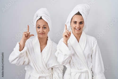 Middle age woman and daughter wearing white bathrobe and towel pointing finger up with successful idea. exited and happy. number one.