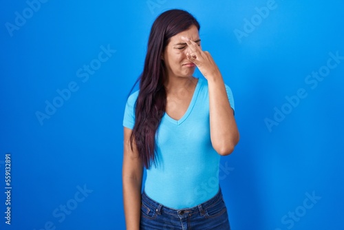 Young hispanic woman standing over blue background smelling something stinky and disgusting, intolerable smell, holding breath with fingers on nose. bad smell