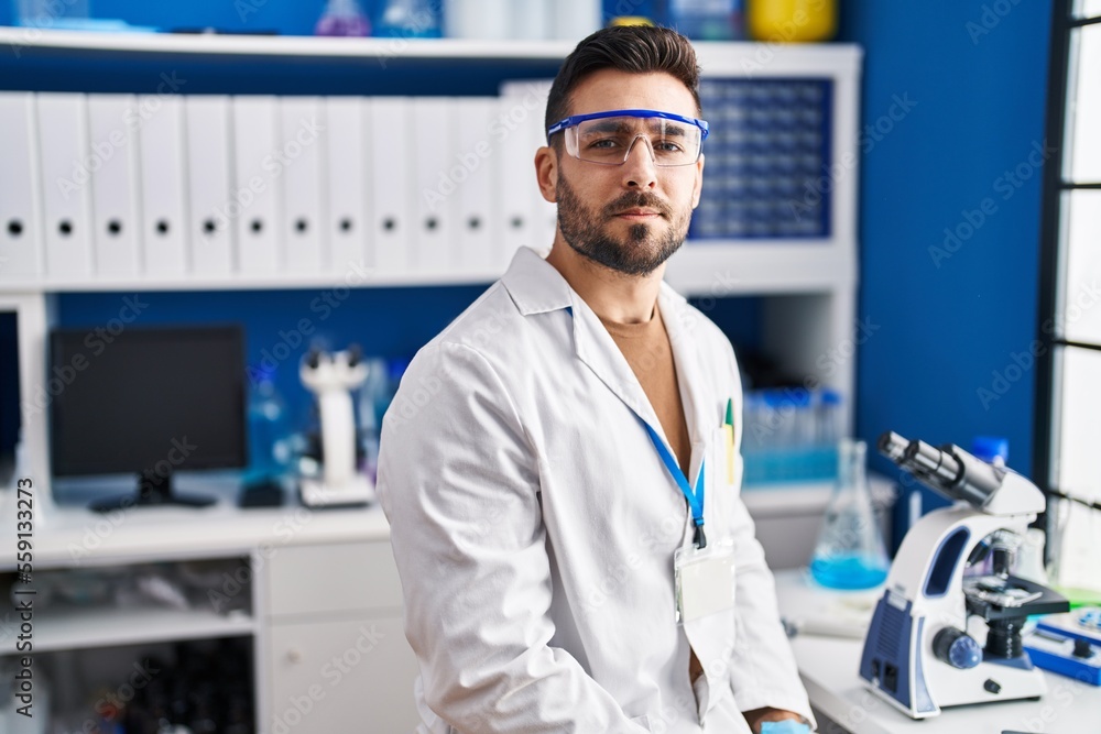 Young hispanic man wearing scientist uniform with serious expression at laboratory Young hispanic man wearing scientist uniform with serious expression at laboratory