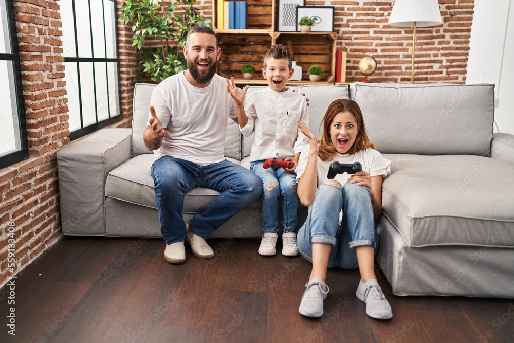 Family of three playing video game sitting on the sofa celebrating victory with happy smile and winner expression with raised hands