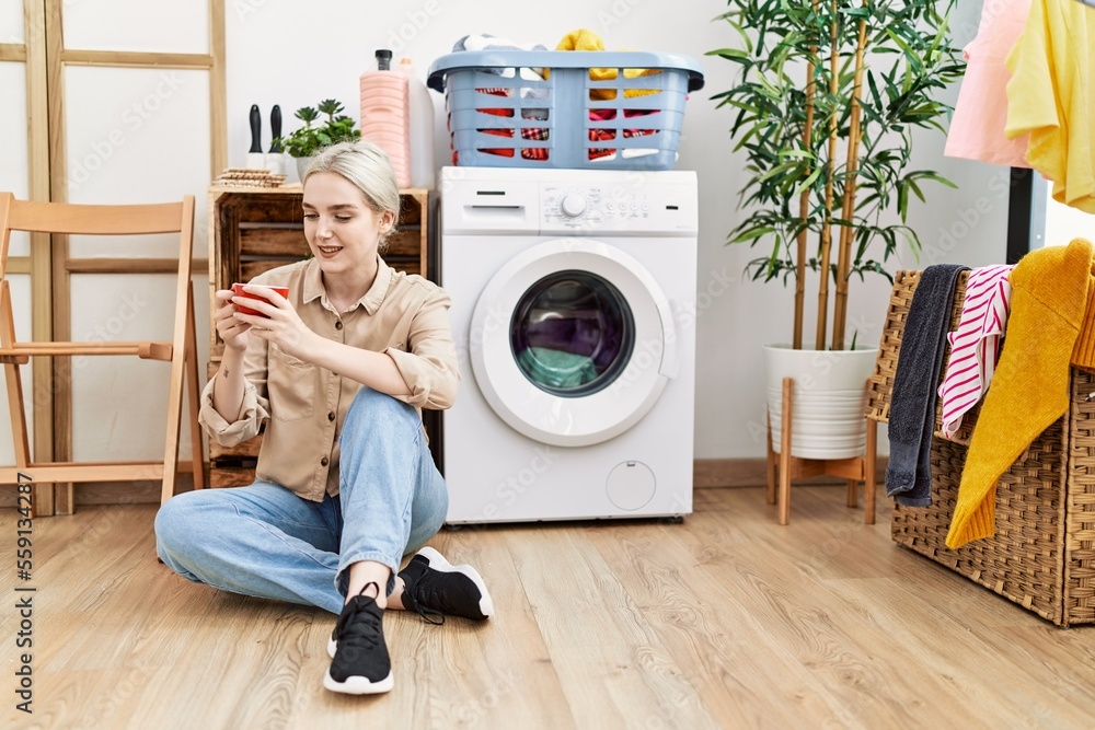 Young caucasian woman drinking coffee waiting for washing machine at laundry room Young caucasian woman drinking coffee waiting for washing machine at laundry room