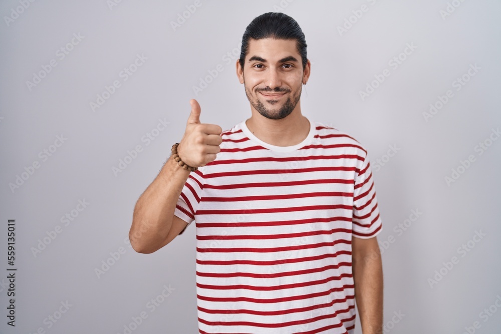 Hispanic man with long hair standing over isolated background doing happy thumbs up gesture with hand. approving expression looking at the camera showing success. Hispanic man with long hair standing over isolated background doing happy thumbs up gesture with hand. approving expression looking at the camera showing success.