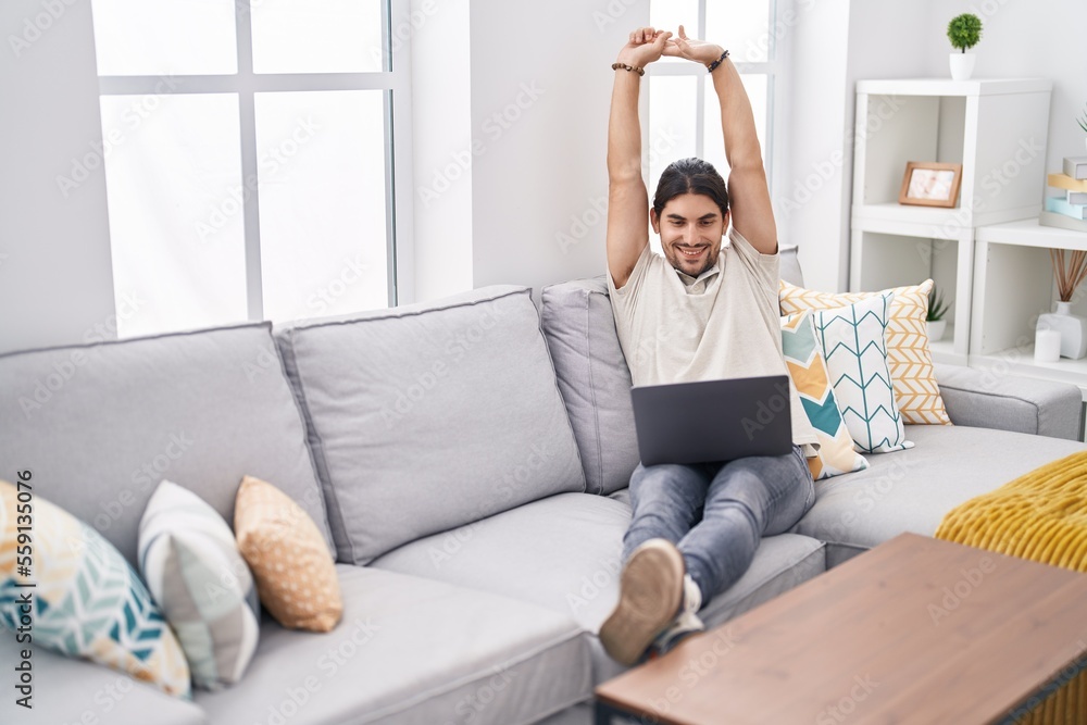 Young hispanic man using laptop stretching arms at home Young hispanic man using laptop stretching arms at home