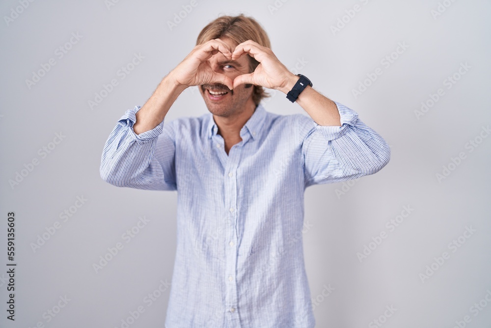 Caucasian man with mustache standing over white background doing heart shape with hand and fingers smiling looking through sign Caucasian man with mustache standing over white background doing heart shape with hand and fingers smiling looking through sign