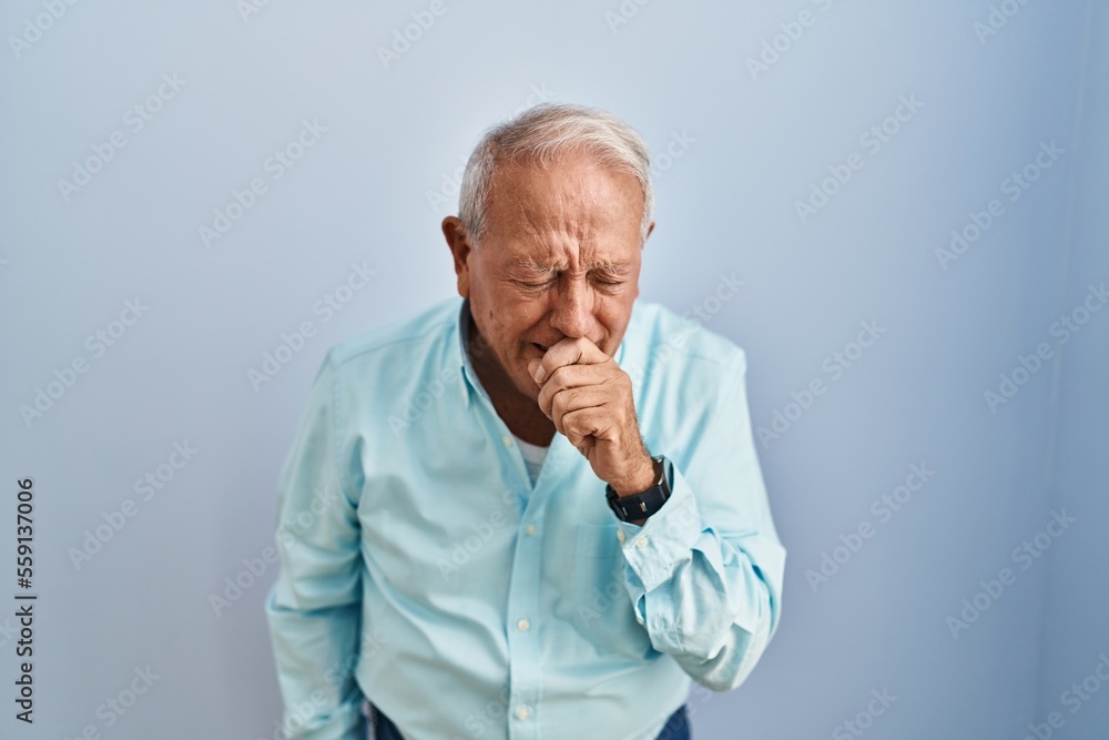 Senior man with grey hair standing over blue background feeling unwell and coughing as symptom for cold or bronchitis. health care concept.