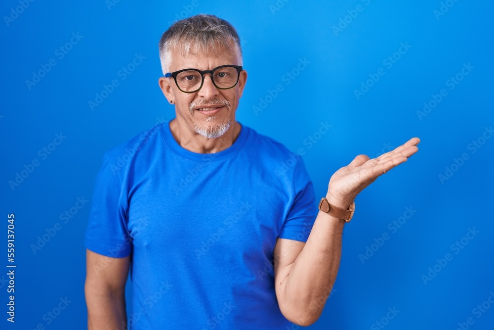 Hispanic man with grey hair standing over blue background smiling cheerful presenting and pointing with palm of hand looking at the camera. Hispanic man with grey hair standing over blue background smiling cheerful presenting and pointing with palm of hand looking at the camera.