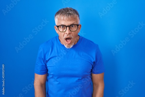 Hispanic man with grey hair standing over blue background afraid and shocked with surprise and amazed expression, fear and excited face. © Krakenimages.com