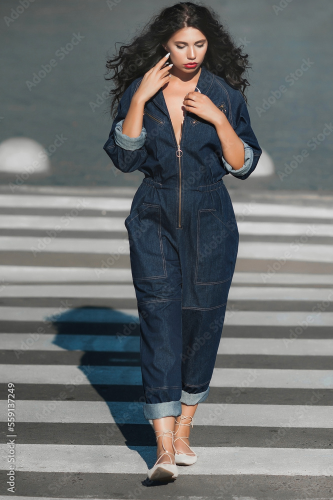 A dark-haired girl in denim overalls; a beautiful girl walks along the road through a pedestrian crossing; red lipstick on her lips