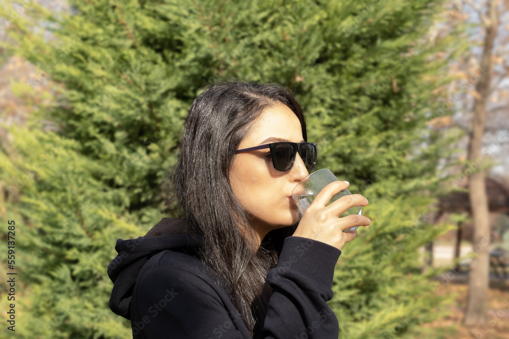 Holding glass, portrait of beautiful woman holding glass. Drinking still clean water. Sunny autumn day. Wearing sunglasses. Concept of health, walking outdoor, jogging, thirsty girl. Copy space. Holding glass, portrait of beautiful woman holding glass. Drinking still clean water. Sunny autumn day. Wearing sunglasses. Concept of health, walking outdoor, jogging, thirsty girl. Copy space.