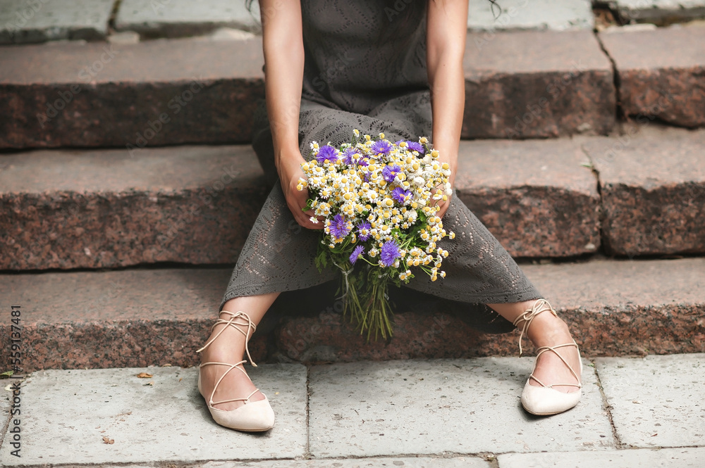 the girl sits on the granite steps and holds flowers in her hands; dressed in a light gray dress