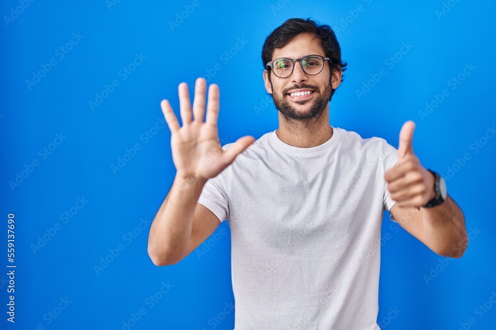 Handsome latin man standing over blue background showing and pointing up with fingers number six while smiling confident and happy. Handsome latin man standing over blue background showing and pointing up with fingers number six while smiling confident and happy.