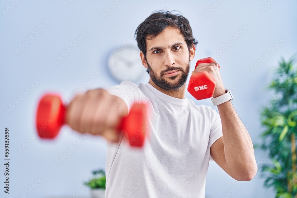 Young hispanic man smiling confident using dumbbells training at home