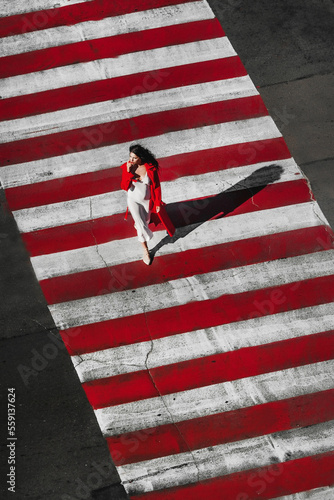 The dark-haired girl is dressed in extravagant evening clothes; a beautiful girl walks along the road through a pedestrian crossing; red lipstick on her lips