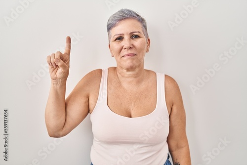 Middle age caucasian woman standing over white background showing and pointing up with finger number one while smiling confident and happy.