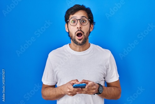 Handsome latin man using smartphone typing message afraid and shocked with surprise and amazed expression, fear and excited face.