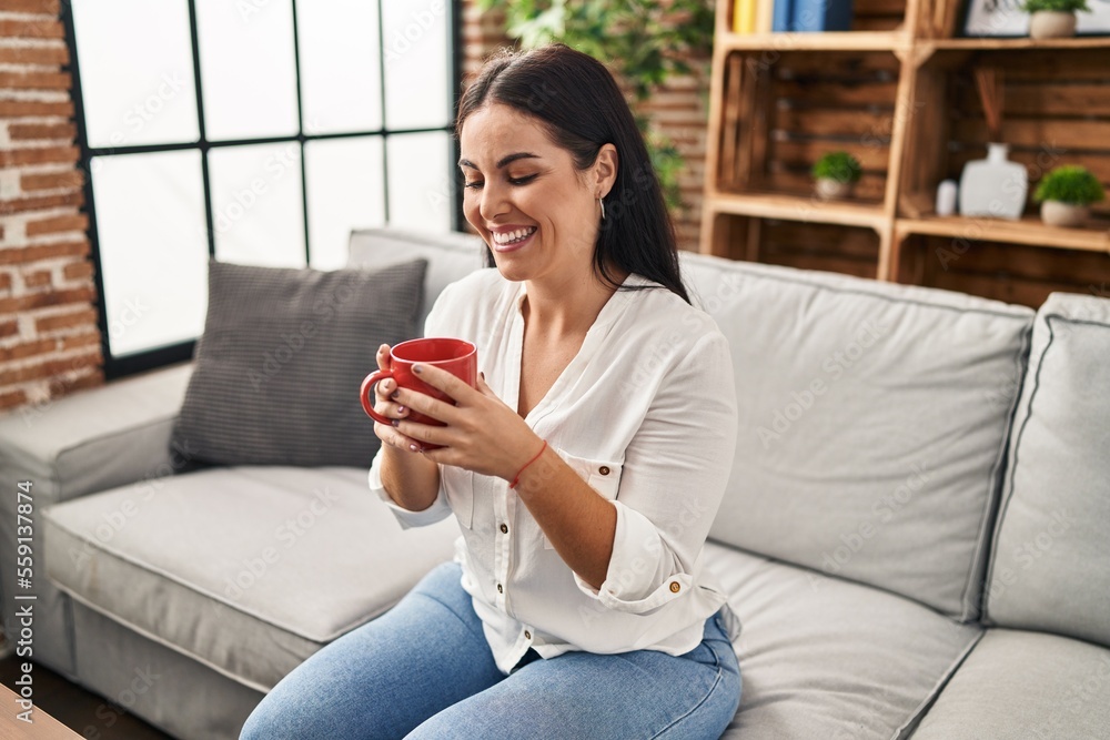 Young beautiful hispanic woman drinking coffee sitting on sofa at home Young beautiful hispanic woman drinking coffee sitting on sofa at home