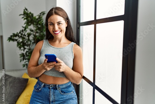 Young beautiful hispanic woman using smartphone standing at home