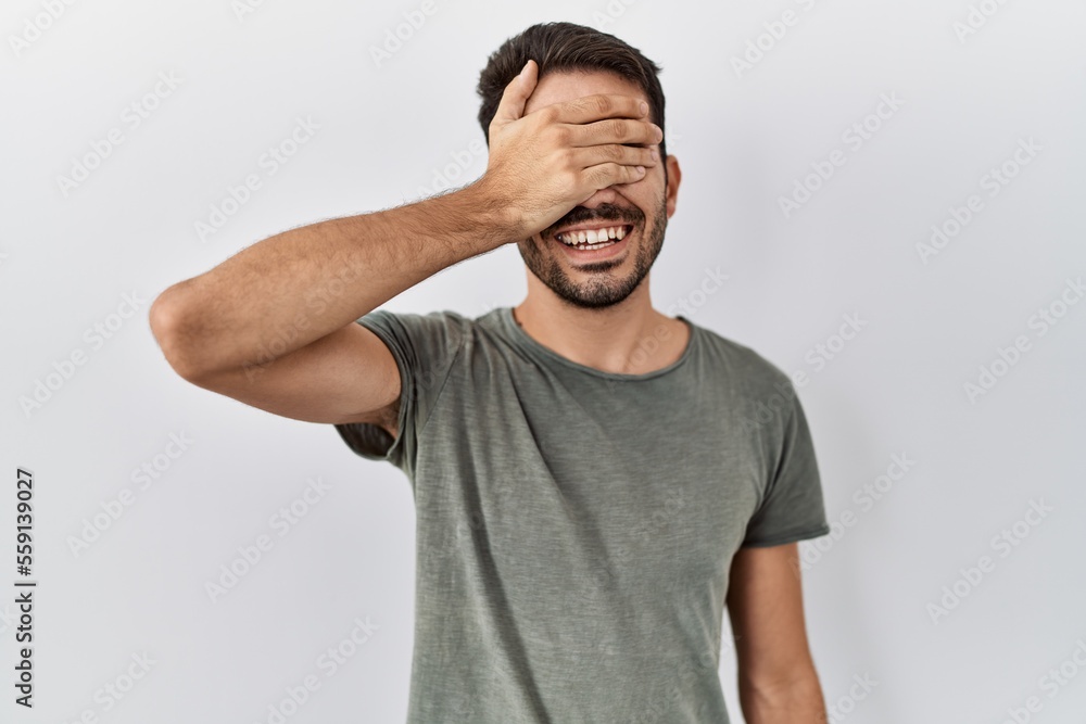 Young hispanic man with beard wearing casual t shirt over white background smiling and laughing with hand on face covering eyes for surprise. blind concept. Young hispanic man with beard wearing casual t shirt over white background smiling and laughing with hand on face covering eyes for surprise. blind concept.