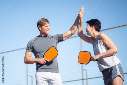 Laughing couple playing pickleball game, hitting pickleball yellow ball with paddle, outdoor sport leisure activity, celebrating victory .