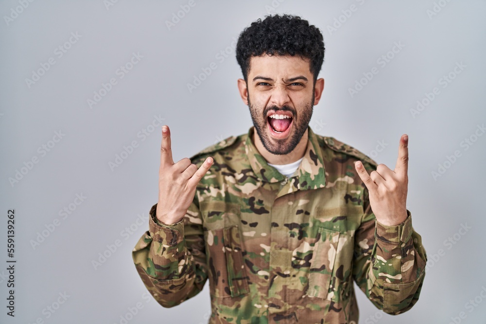 Arab man wearing camouflage army uniform shouting with crazy expression doing rock symbol with hands up. music star. heavy music concept.