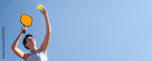 Pickleball banner over blue sky, woman playing pickleball game, hitting pickleball yellow ball with paddle, outdoor sport leisure activity, copyspace for text.