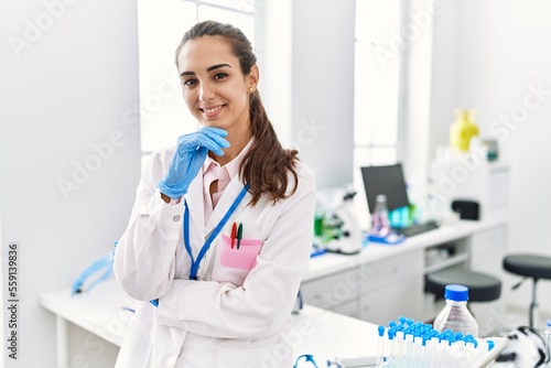 Young hispanic woman wearing scientist uniform standing with arms crossed gesture at laboratory