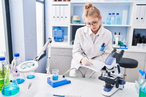 Young blonde woman scientist writing on notebook weighing liquid at laboratory
