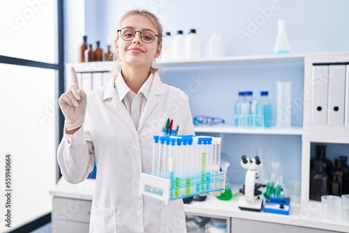 Young caucasian woman working at scientist laboratory holding samples smiling with an idea or question pointing finger with happy face, number one
