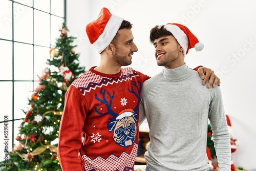 Two hispanic men couple hugging each other standing by christmas tree at home