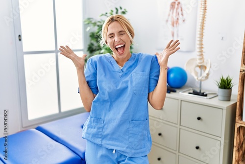 Beautiful blonde physiotherapist woman working at pain recovery clinic celebrating mad and crazy for success with arms raised and closed eyes screaming excited. winner concept