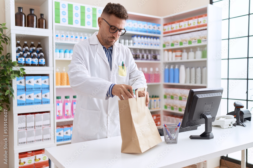 Young hispanic man pharmacist putting medication bottle on shopping bag at pharmacy