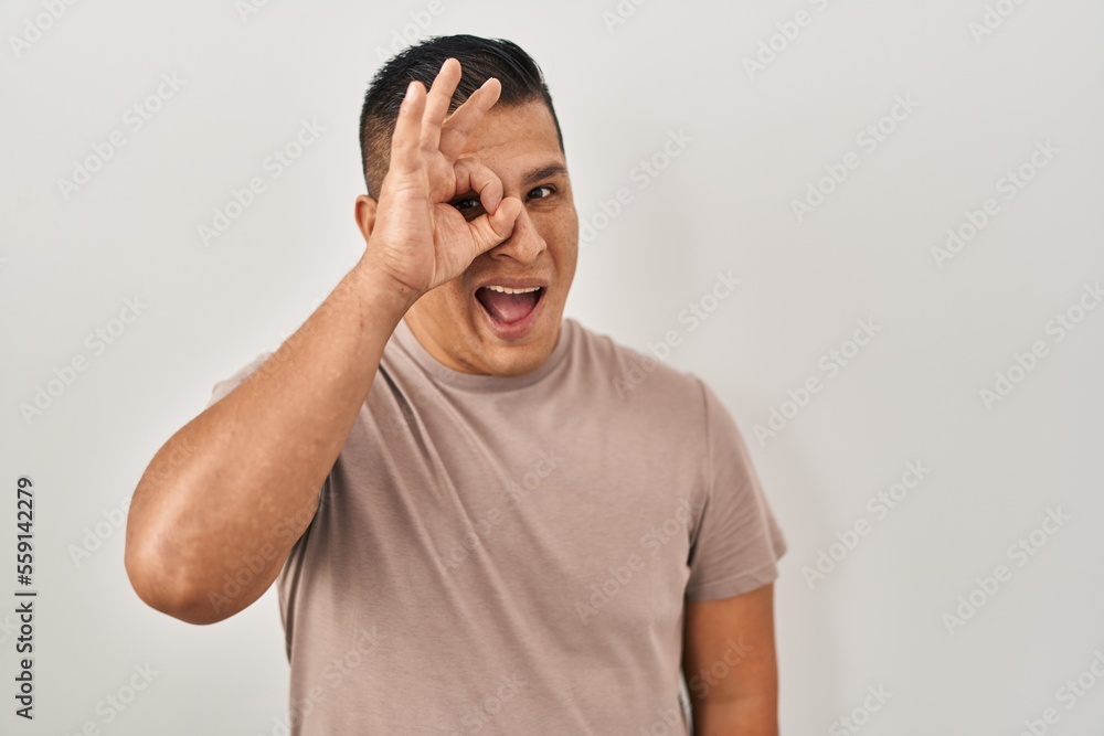 Hispanic young man standing over white background doing ok gesture with hand smiling, eye looking through fingers with happy face. Hispanic young man standing over white background doing ok gesture with hand smiling, eye looking through fingers with happy face.