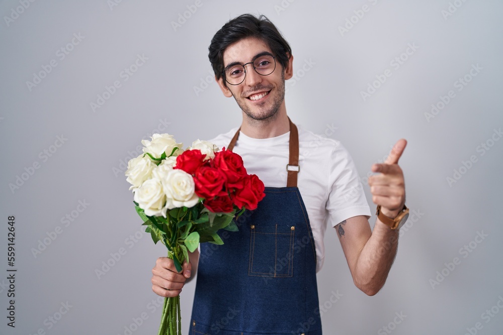 Young hispanic man holding bouquet of white and red roses pointing fingers to camera with happy and funny face. good energy and vibes. Young hispanic man holding bouquet of white and red roses pointing fingers to camera with happy and funny face. good energy and vibes.