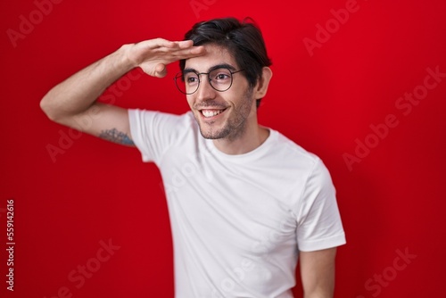 Young hispanic man standing over red background very happy and smiling looking far away with hand over head. searching concept.