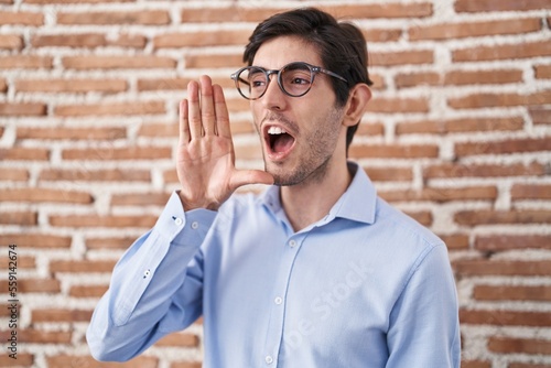 Young hispanic man standing over brick wall background shouting and screaming loud to side with hand on mouth. communication concept.
