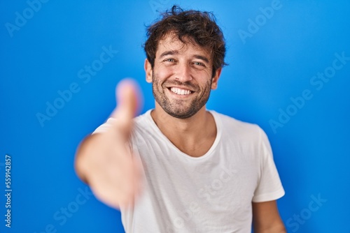 Hispanic young man standing over blue background smiling friendly offering handshake as greeting and welcoming. successful business.