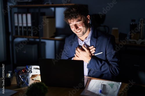 Hispanic young man working at the office at night smiling with hands on chest with closed eyes and grateful gesture on face. health concept.